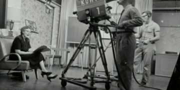 grayscale photography of man standing near studio camera and woman sitting while holding book