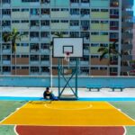 man sitting below basketball hoop near the building during daytime