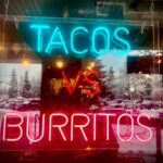 Eye-catching neon sign highlighting tacos vs burritos debate in a shop window with snowy reflections.