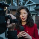 Asian woman reporting news on camera outdoors, wearing red coat.