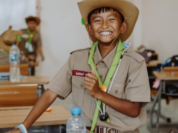 Cheerful boy scout in a classroom setting, wearing uniform and hat, smiling widely.