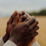 A close-up of two people of different ethnicities holding hands outdoors, symbolizing unity and support.