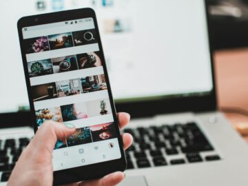 A person uses a smartphone to browse social media in front of a laptop, emphasizing modern technology and lifestyle.