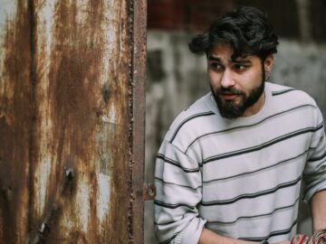 Man in striped sweater stands thoughtfully by a rusty doorway, striking a contemplative pose.