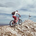 An elderly man biking uphill on a rocky mountain trail under a cloudy blue sky.