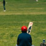 Man wearing red cap sitting on grass field holding a cricket bat, blurred player in background.