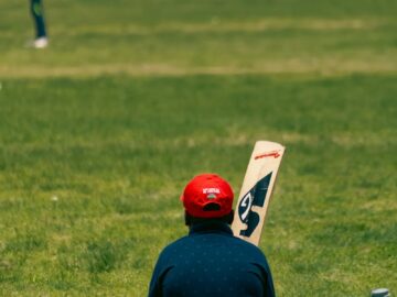 Man wearing red cap sitting on grass field holding a cricket bat, blurred player in background.