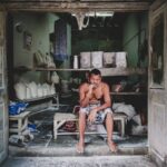Shirtless man sits smoking in an old, rustic storefront filled with goods.