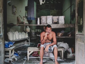 Shirtless man sits smoking in an old, rustic storefront filled with goods.