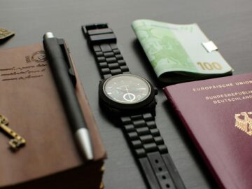 A neatly arranged flat lay depicting essential travel items such as a German passport, wristwatch, and euro bills on a desk.