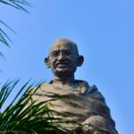 Statue of Mahatma Gandhi against a blue sky backdrop with foliage framing.