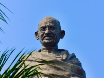 Statue of Mahatma Gandhi against a blue sky backdrop with foliage framing.