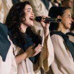 Woman singing with a choir, passionately holding a microphone. Captured in mid-performance with focused expression.