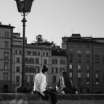 Two women sit on a wall in Tuscany, Italy, with historical buildings in the background.