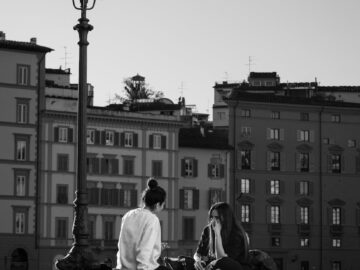 Two women sit on a wall in Tuscany, Italy, with historical buildings in the background.