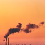 Orange sky at sunset with industrial smokestacks emitting smoke over water.