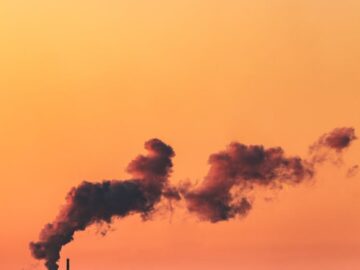 Orange sky at sunset with industrial smokestacks emitting smoke over water.
