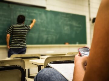 Student texting in a classroom while teacher is writing on the blackboard.