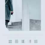 A man in a coat walks up a minimalist white staircase holding a camera.