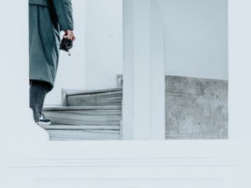 A man in a coat walks up a minimalist white staircase holding a camera.