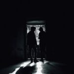 Silhouetted figures stand in a dim room with sunlight streaming through a doorway.