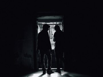 Silhouetted figures stand in a dim room with sunlight streaming through a doorway.