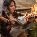 A woman reads a newspaper while it's on fire, creating a dramatic scene with smoke outdoors.