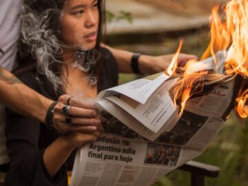 A woman reads a newspaper while it's on fire, creating a dramatic scene with smoke outdoors.