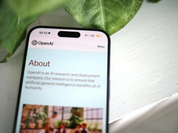 Close-up shot of a smartphone screen showing the OpenAI website with greenery in the background.