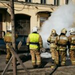 Firefighters responding to a car fire on a street in St. Petersburg, Russia, with smoke and safety gear in view.