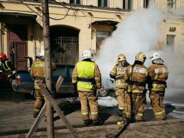 Firefighters responding to a car fire on a street in St. Petersburg, Russia, with smoke and safety gear in view.