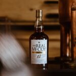 A bottle of The Real McCoy 12-year aged rum on a wooden shelf with a copper still in the background.
