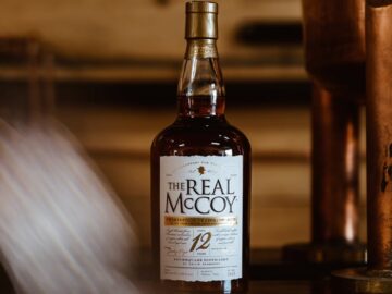 A bottle of The Real McCoy 12-year aged rum on a wooden shelf with a copper still in the background.