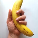 Close-up of a hand holding a ripe yellow banana against a white backdrop, showcasing freshness and tropical vibes.
