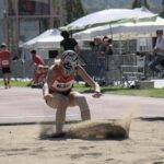 Female athlete captured mid-landing in the long jump event during a sunny sports meet.