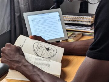 A man studying with a book and tablet at a desk indoors, showcasing multitasking and modern study habits.
