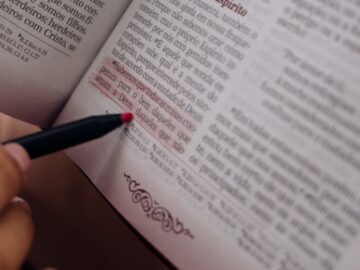 A hand with red pen highlights text in an open book. Perfect for education themes.