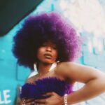 A fashionable woman with afro hair poses confidently in a vibrant purple outfit against a colorful outdoor backdrop.