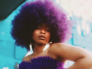 A fashionable woman with afro hair poses confidently in a vibrant purple outfit against a colorful outdoor backdrop.