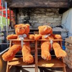 Two plush teddy bears seated on wooden chairs in front of a rustic grill setting.