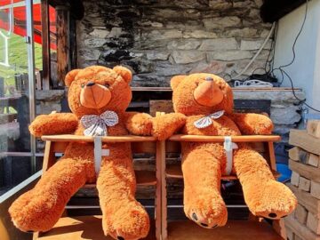 Two plush teddy bears seated on wooden chairs in front of a rustic grill setting.