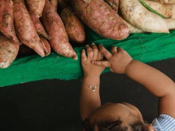 A young child curiously reaches for fresh sweet potatoes at an outdoor market stall.