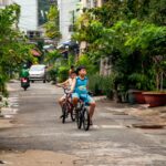 Two boys enjoy a bike ride on an urban residential street surrounded by greenery.
