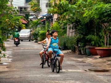 Two boys enjoy a bike ride on an urban residential street surrounded by greenery.