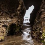 Silhouetted couple exploring a rocky archway at Piha Beach, Auckland, New Zealand.