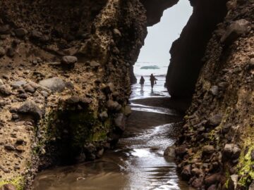 Silhouetted couple exploring a rocky archway at Piha Beach, Auckland, New Zealand.