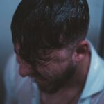 A man stands in the shower with water cascading over him, reflecting a moody, blue tone.