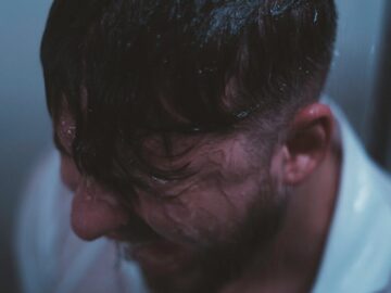 A man stands in the shower with water cascading over him, reflecting a moody, blue tone.