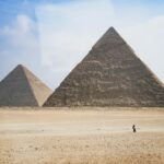 Wide shot of the Pyramids of Giza at daytime with a distant solitary figure.
