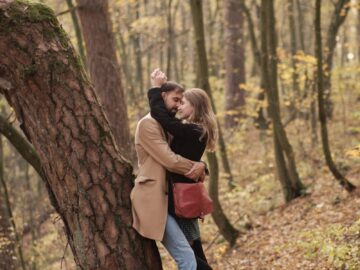 A couple lovingly embraces against a tree in a serene autumn forest setting.